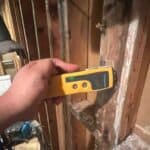 A hand holds a yellow moisture meter against a wooden wall stud