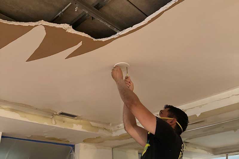 A person wearing a black t-shirt and a face mask uses a screwdriver to work on a white circular fixture in a damaged ceiling