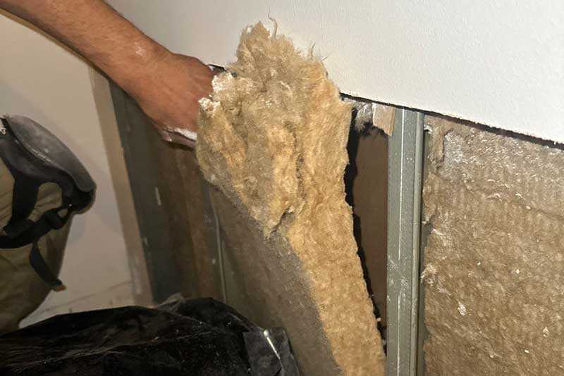 Hand removing damaged insulation from an exposed wall, revealing internal structure and potential damage.