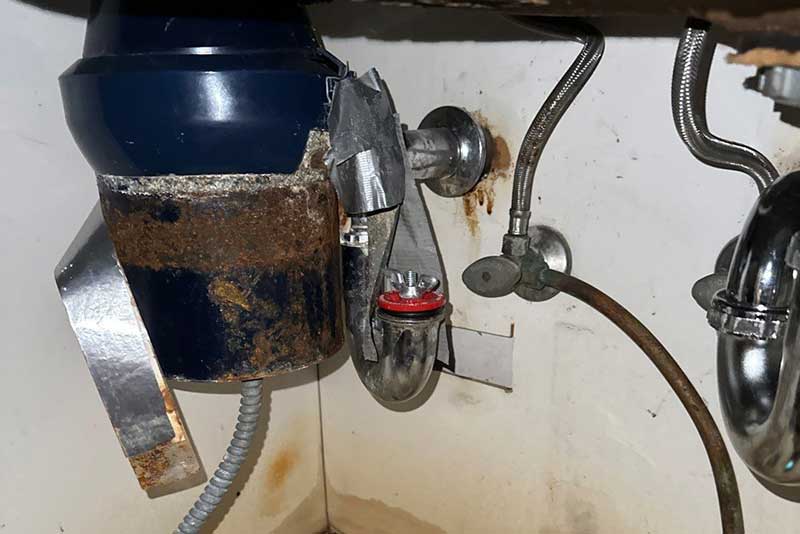 Rusted garbage disposal and corroded plumbing under a kitchen sink, showing water damage.
