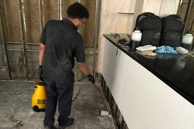 Worker applies pest treatment with a sprayer inside an unfinished building.